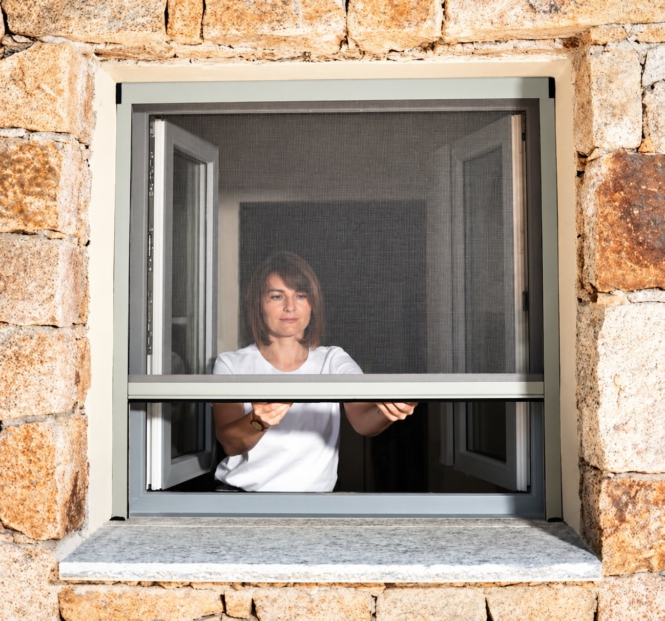 Une femme en t-shirt blanc tire vers le bas une moustiquaire enroulable installée sur une fenêtre dans un mur en pierres apparentes.