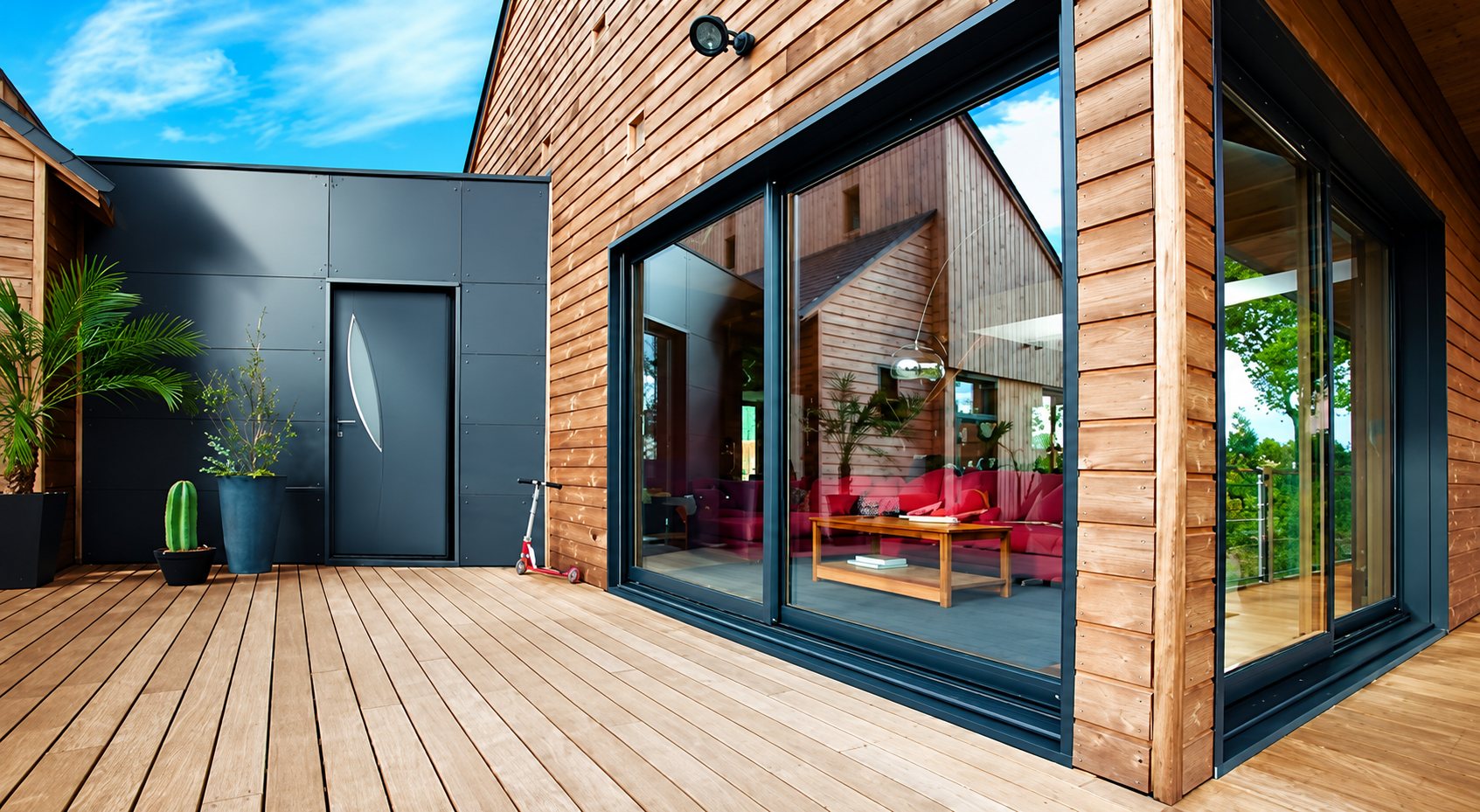 Façade d'une maison moderne en bardage bois avec grande baie vitrée coulissante anthracite, porte d'entrée grise et terrasse en bois.